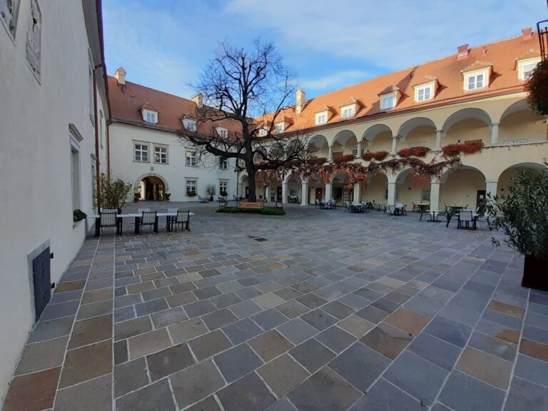 Großer, gepflasterter Innenhof mit Arkadengängen und einem Baum in der Mitte.