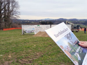 Nahaufnahme einer Informationsbroschüre zum Wohnprojekt Bartholomä vor dem Infoplakat und Bagger beim Spatenstich.
