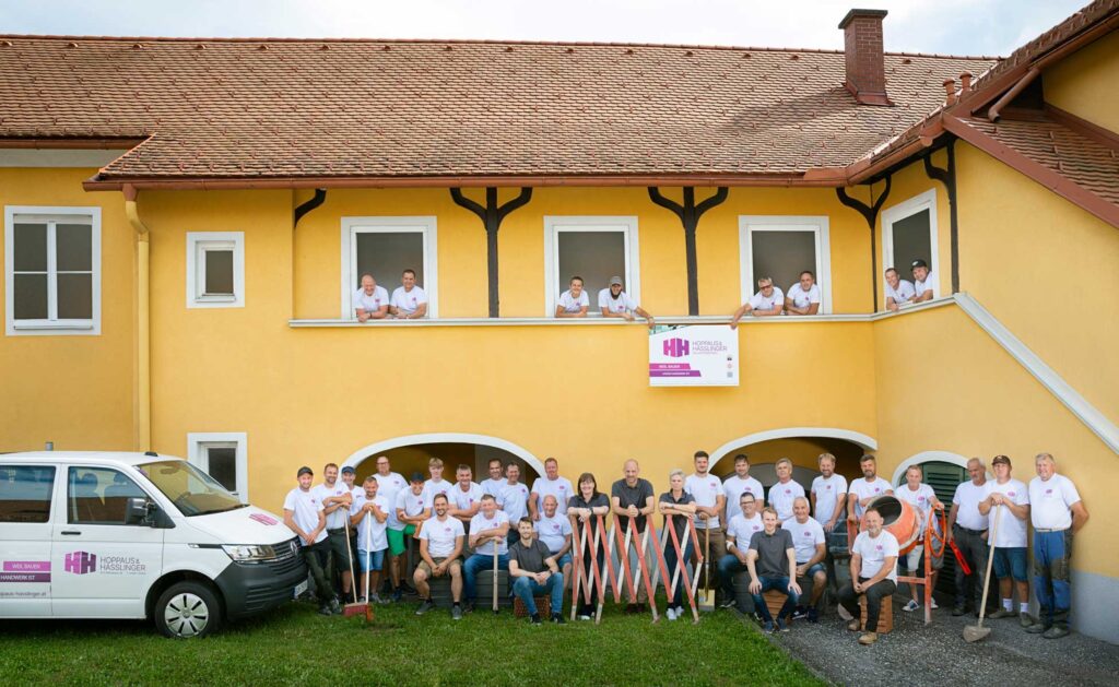 Gruppenfoto des gesamten Teams von Hoppaus & Hasslinger vor dem gelben Firmengebäude