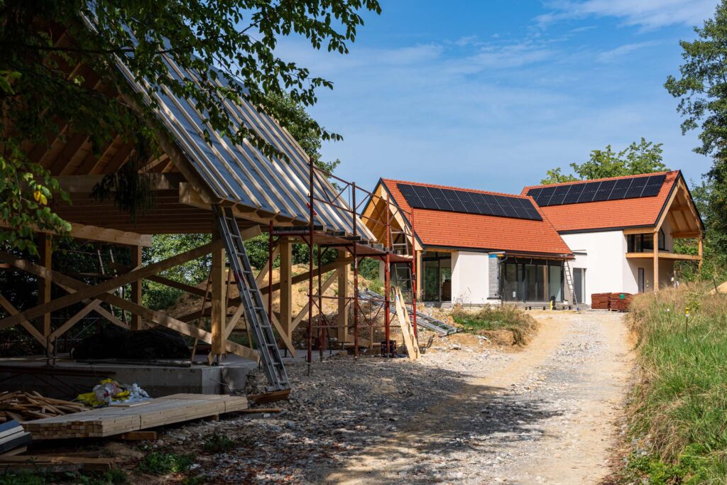 Baustellenzufahrt zwischen einer offenen Holzkonstruktion und einem Einfamilienhaus mit roten Ziegeldächern und Photovoltaikmodulen.