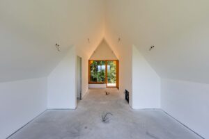 Dachraum im Innenausbau mit weißen Dachschrägen, sichtbaren Elektroanschlüssen und einem Fenster mit Glastür am Raumende.