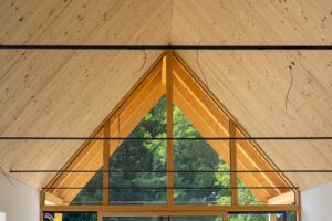 Dreieckige Giebelverglasung mit Holzrahmen unter einer sichtbaren Holzdecke, davor horizontale schwarze Zugstangen.