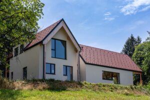 Einfamilienhaus mit heller Putzfassade, roten Ziegeldächern und großem Giebelfenster auf einem begrünten Hanggrundstück.