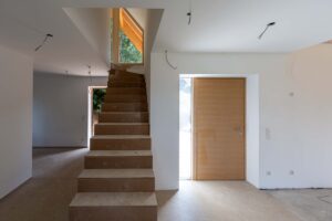 Eingangsbereich im Innenausbau mit noch unverkleideter Treppe, Holzeingangstür und hohem Fenster am Treppenaufgang.