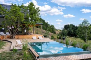 Pool mit angrenzender Holzterrasse und Liegestühlen vor einem Wohnhaus in einem begrünten Hanggrundstück.
