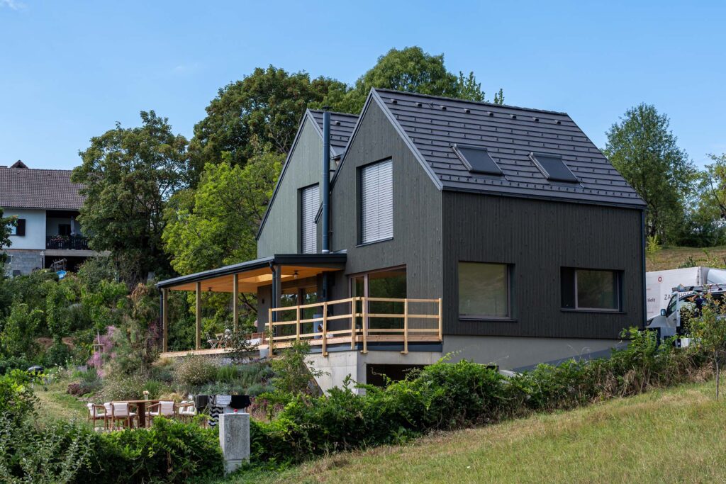 Wohnhaus in Hanglage mit dunkler Holzfassade, zwei versetzten Satteldächern und seitlich überdachter Terrasse.