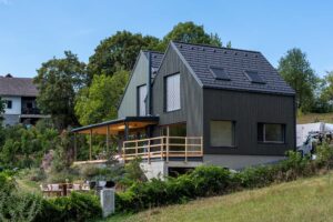 Wohnhaus in Hanglage mit dunkler Holzfassade, zwei versetzten Satteldächern und seitlich überdachter Terrasse.