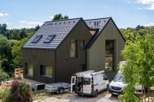 Wohnhaus mit dunkler Holzfassade, versetzten Satteldächern und Dachfenstern, davor Handwerkerfahrzeuge an der Zufahrt.